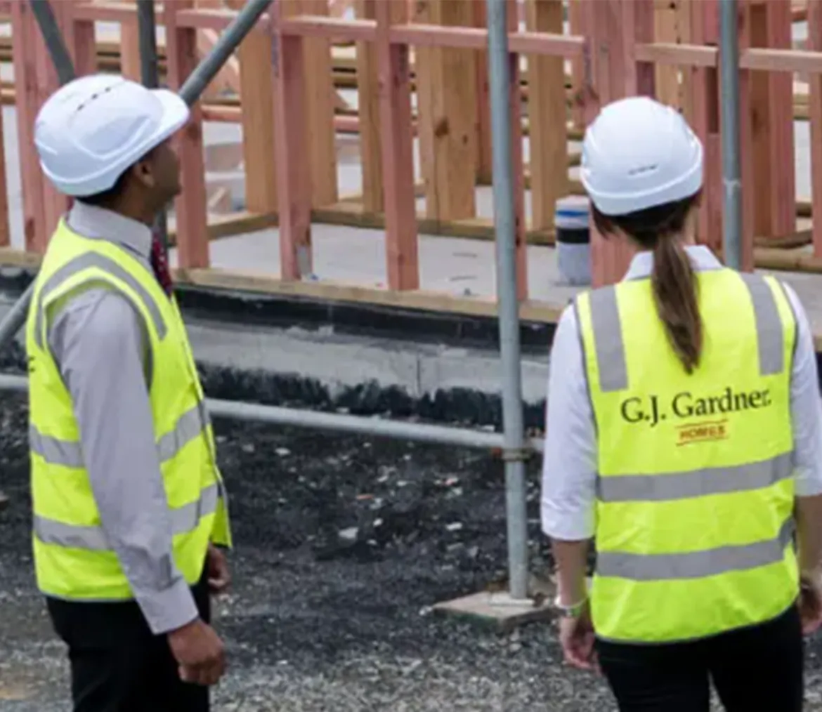 Two G.J. Gardner workers wearing hard hats and high-visibility vests beside timber framing