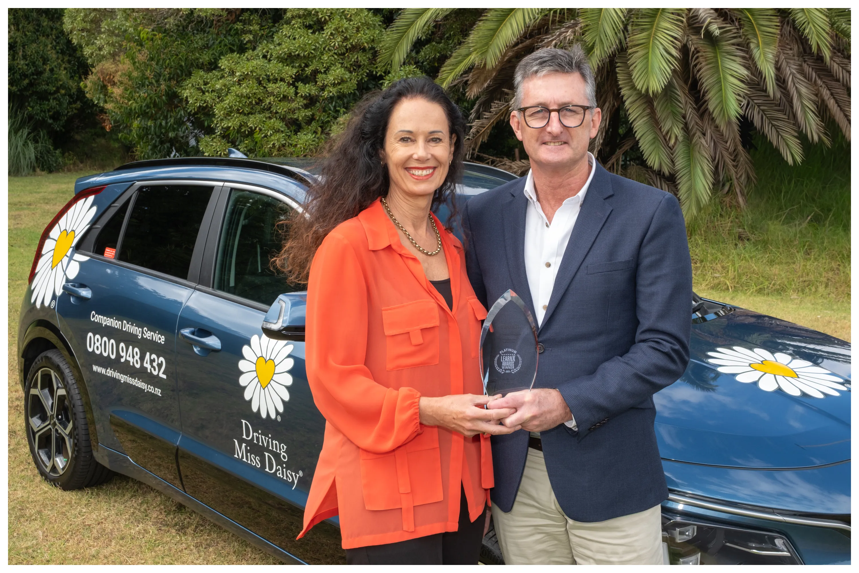 Two people holding a LearnX Platinum award beside a Driving Miss Daisy vehicle