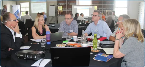 Alternative Board participants discussing business around a table with laptops, notebooks and refreshments